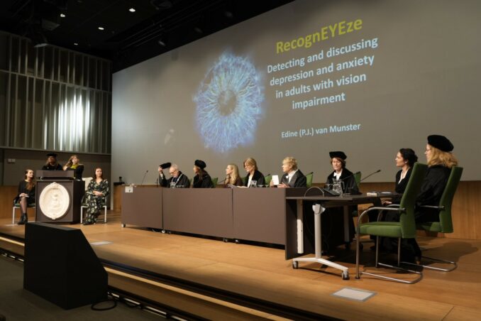 Foto van promovendus Edine van Munster met haar paranimfen aan haar zijde op het podium, klaar om te starten met het verdedigen van haar proefschrift. Rechts op het podium de leden van de commissie.
