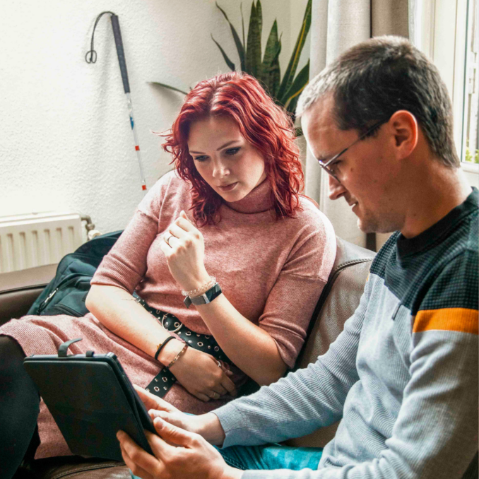 Website – cl en begl met tablet – 1000×1000 Een cliënt en een begeleider zitten samen op een bank en bekijken informatie op een tablet.